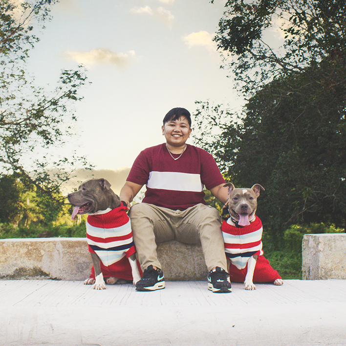 Two dogs wearing red and white striped sweaters sit on either side of young, smiling person framed by trees and a blue sky