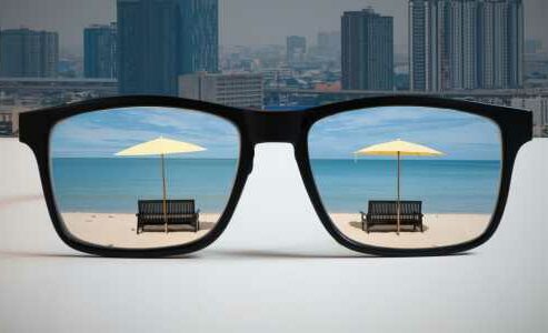 A reflection of a beach appears in a pair of sunglasses. Behind the sunglasses, there is a cityscape.